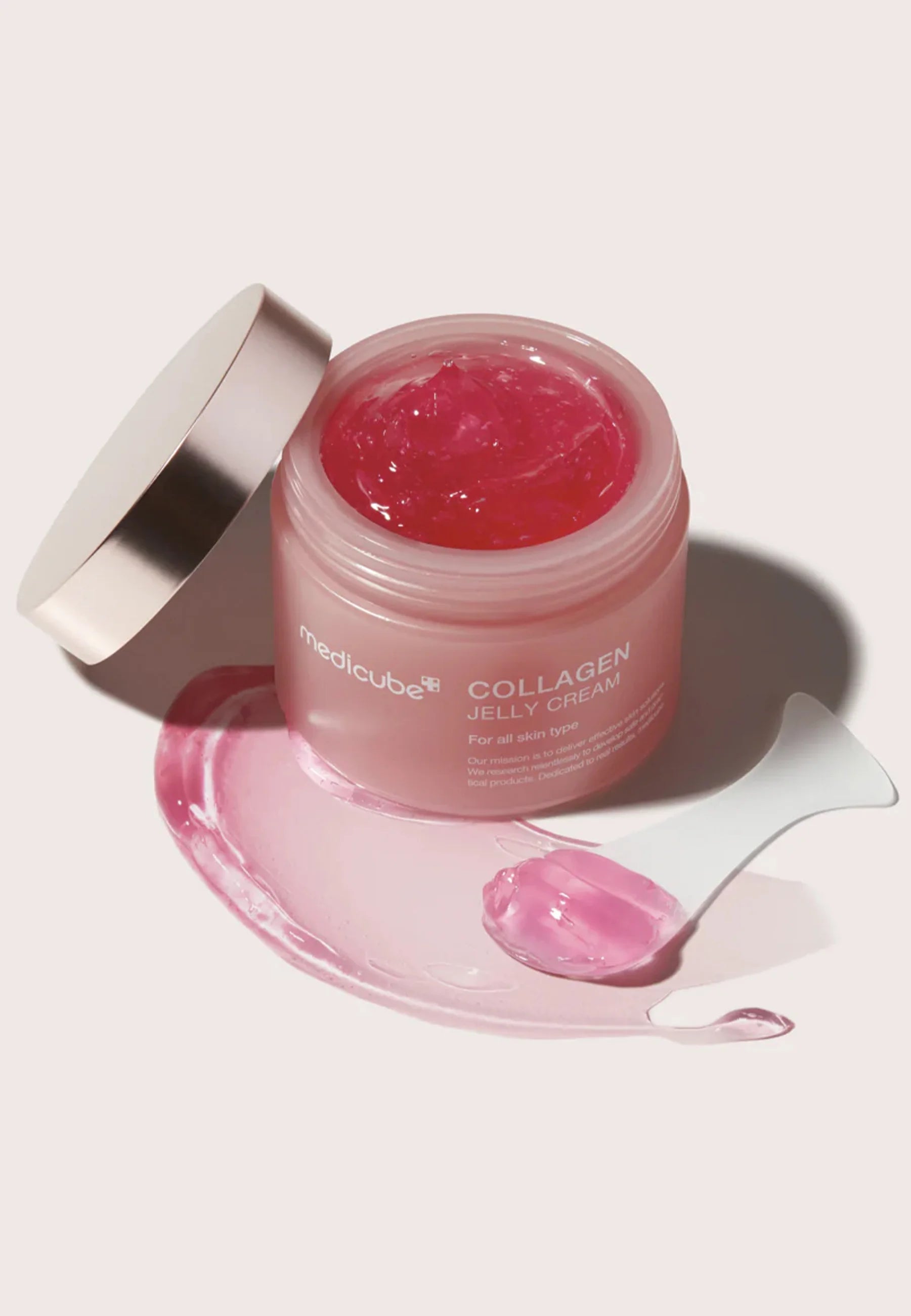 Öppen burk med Medicube Collagen Jelly Cream och rosa gel utsmetad bredvid med spatel på ljus bakgrund.